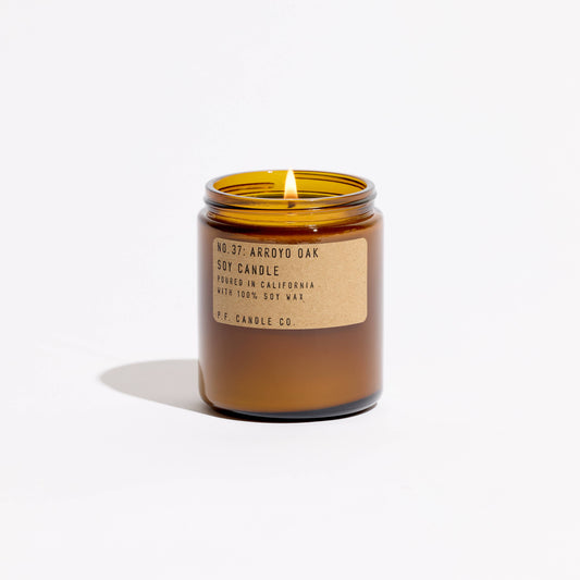 Arroyo Oak soy candle lit with lid off, amber glass jar with kraft label on a white background