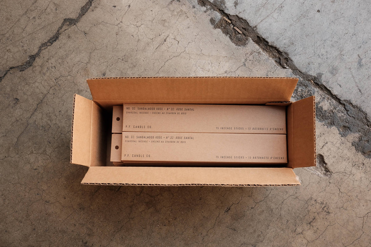 Open cardboard shipping box holding two slim kraft incense boxes, seen from above on concrete