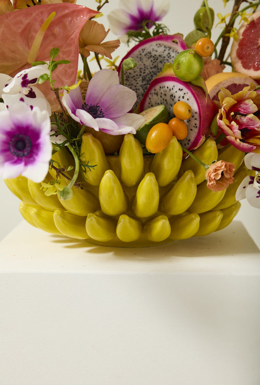Close-up of yellow banana bowl overflowing with dragon fruit, kumquats, grapefruit and anemone flowers