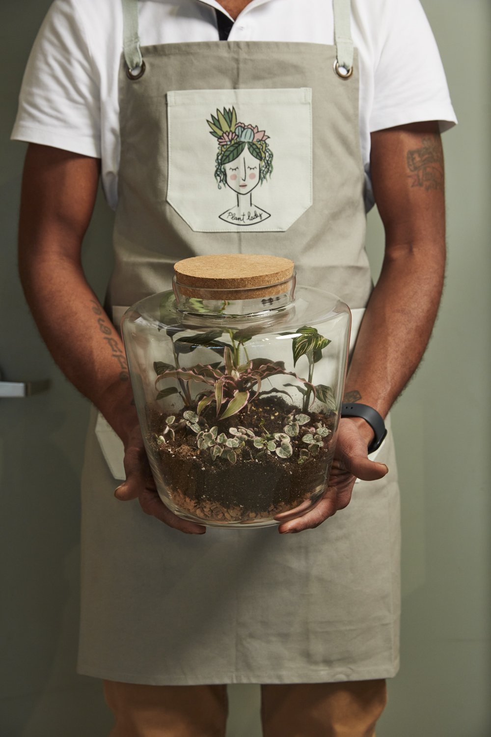 Person in an apron holding the cork-lidded Message terrarium planted with ferns and moss