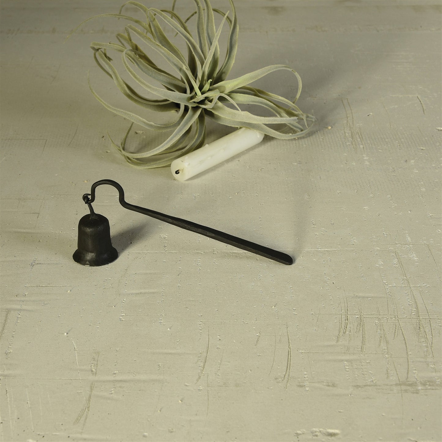black hand-forged iron snuffer on concrete with candle and air plant in background