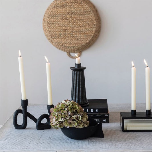 collection of abstract and modern taper holders on table with lit white candles. Rattan round basket hanging on background.