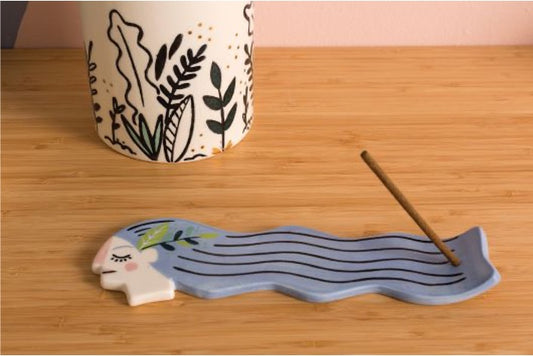 Incense holder in shape of a female head with long hair in blue and incense stick at the end.