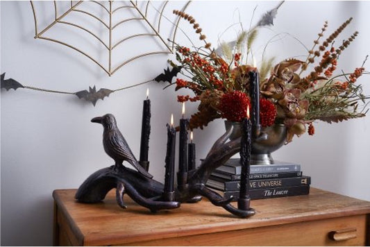 Metal candleholder in dark bronze with lit black candles on wooden table with dried fall foliage. Bat and web wall decor in background.