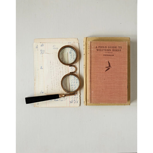 Brass spectacle magnifying glass with black handle next to vintage paper and books