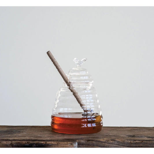 Clear glass honey pot with raw honey and bamboo dipper on wooden table.