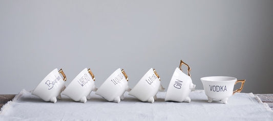 Six tea cups with gold handles tilted on table. Each tea cup has black text with Wine, Vodka, Gin, Tequila, Whiskey, or Bourbon on side.
