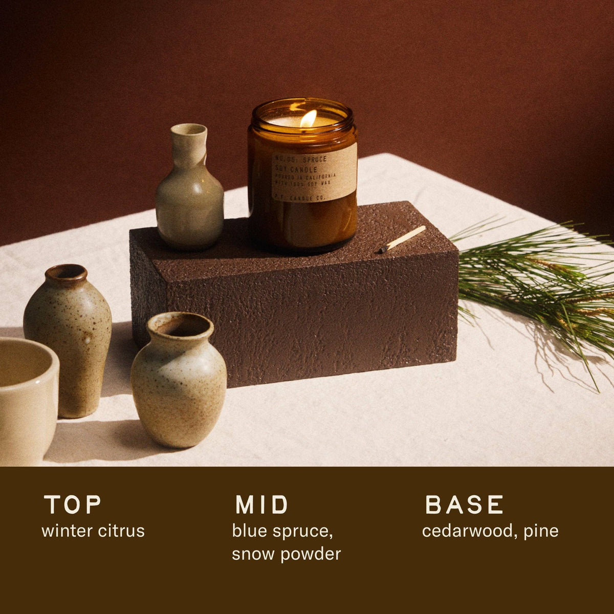A lit amber glass candle jar sits on a dark brown textured block, styled with ceramic vases and pine branches on a cream surface against a warm brown background. Text at the bottom reads 'TOP winter citrus', 'MID blue spruce, snow powder', and 'BASE cedarwood, pine'.