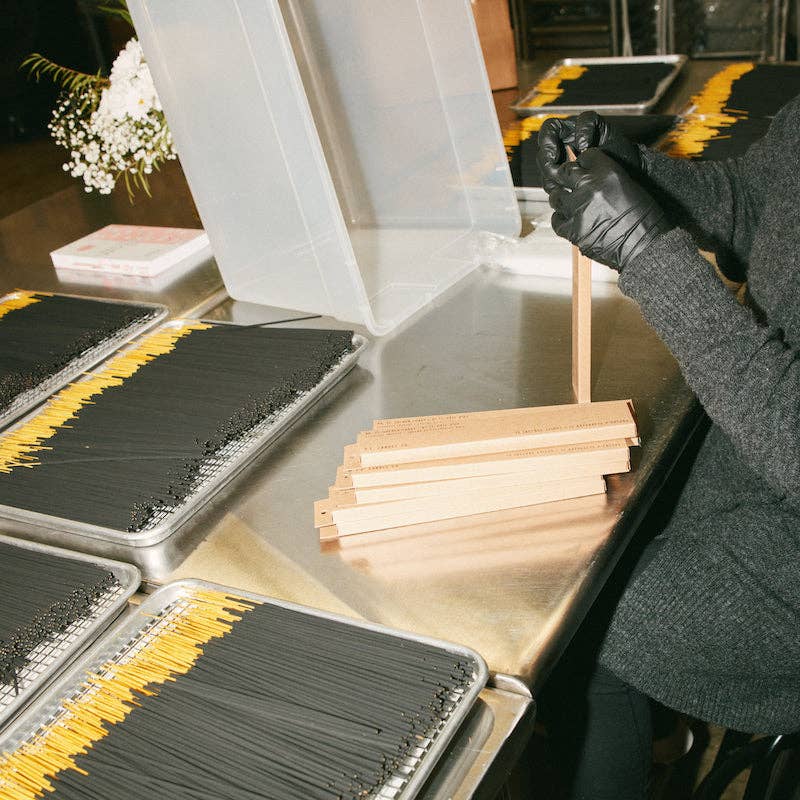 Gloved hands packing incense into kraft boxes at a steel table lined with trays of black incense sticks