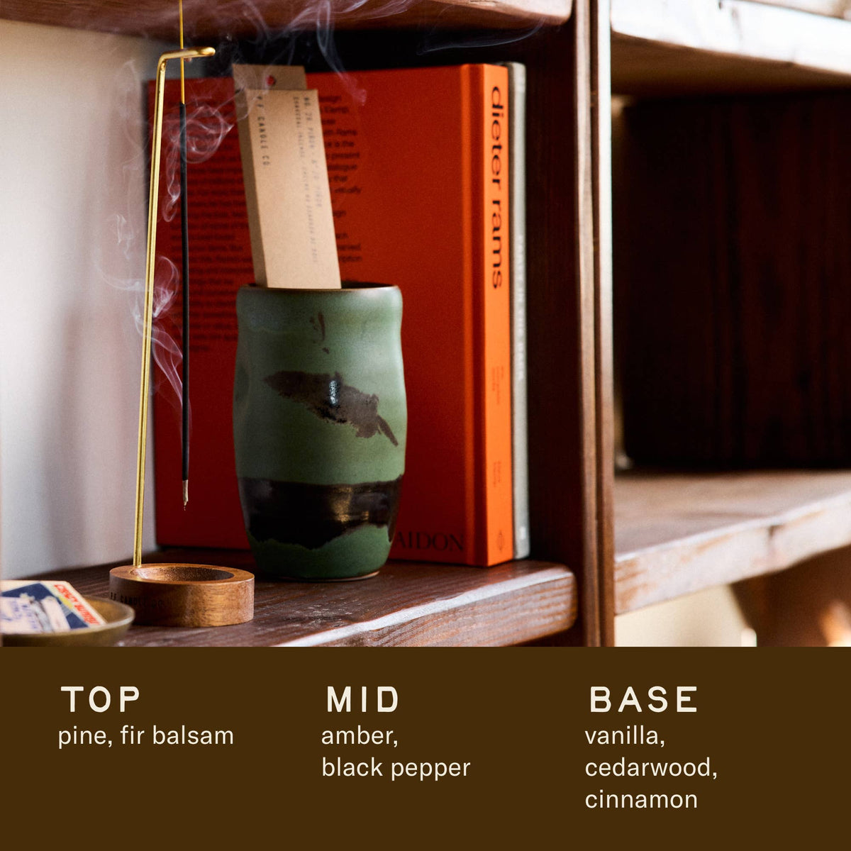 Lit Piñon incense on a shelf with scent notes below: top pine, fir balsam; mid amber, black pepper; base vanilla
