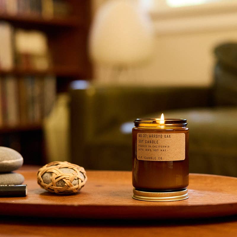 Lit Arroyo Oak candle in amber jar on a wooden tray beside stones, with a green sofa and bookshelf behind