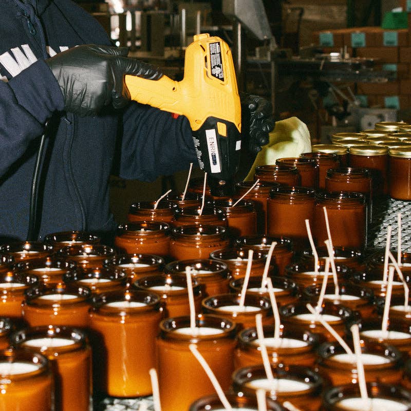 Gloved hands using a glue gun to set wicks in rows of amber candle jars