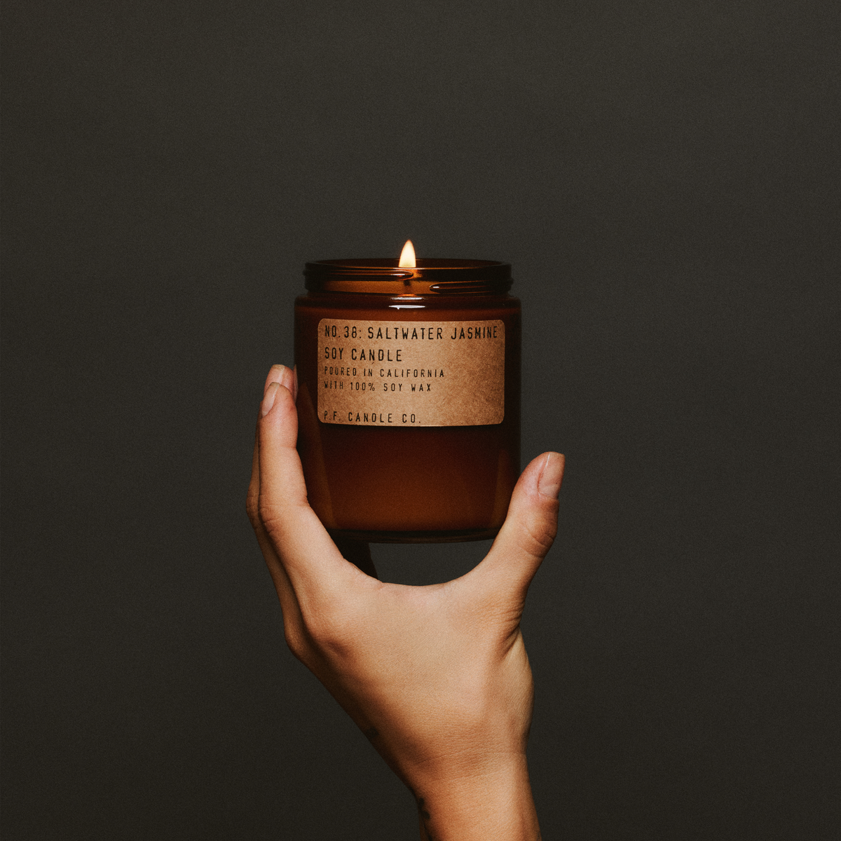 Hand holding up a lit Saltwater Jasmine candle in its amber jar against a dark gray wall