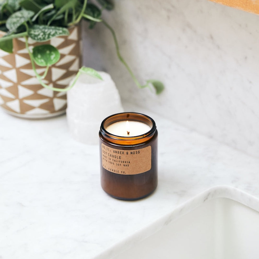 No. 11 Amber & Moss Soy candle in amber jar with craft label and brass lid. Placed on white marble counter.