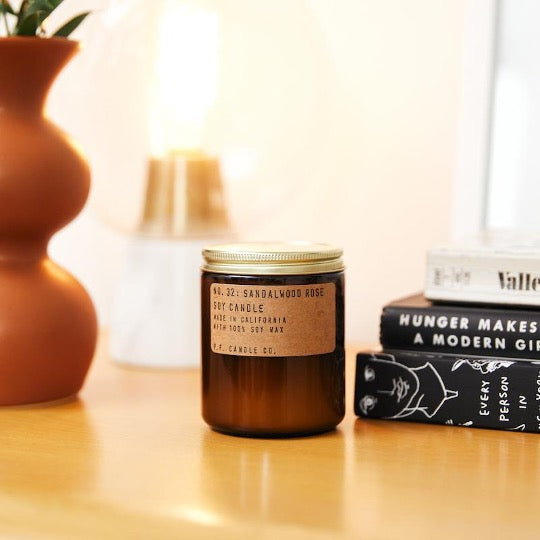 No. 32 Sandalwood Rose Soy candle in amber jar with craft label and brass lid.  On table next to books and vase. 