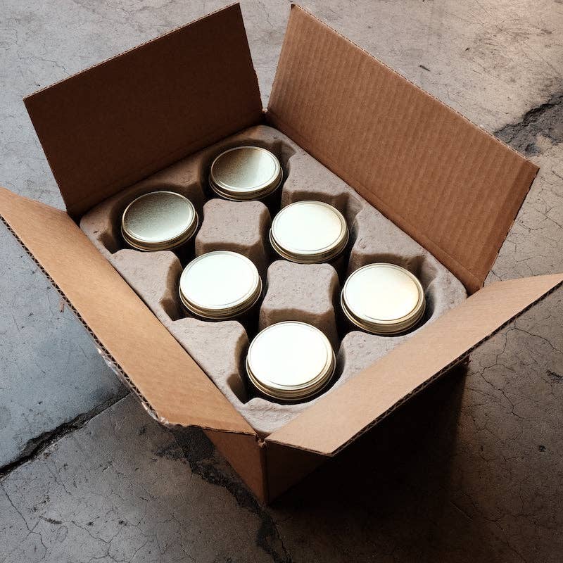Open cardboard shipping box holding six gold-lidded candle jars in a molded pulp insert, seen from above