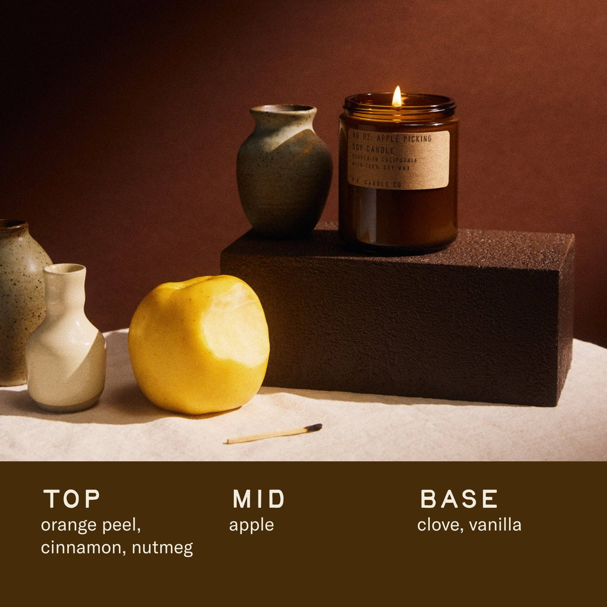 Lit Apple Picking candle on a dark block beside a yellow apple and vases; notes orange peel, apple, clove, vanilla