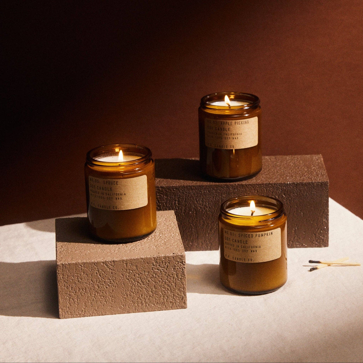 Three lit amber jar candles, Apple Picking, Spruce and Spiced Pumpkin, on textured blocks against rust red