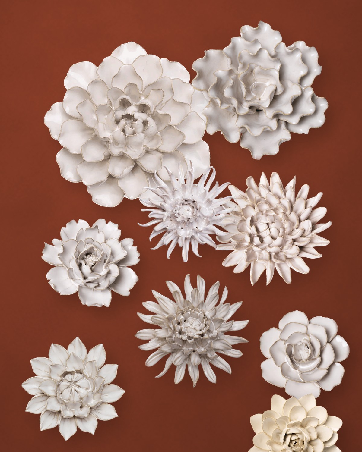 Nine ivory ceramic flowers, including a water lily, sea lettuce, chrysanthemum and spider mum, clustered on a rust-colored plaster wall