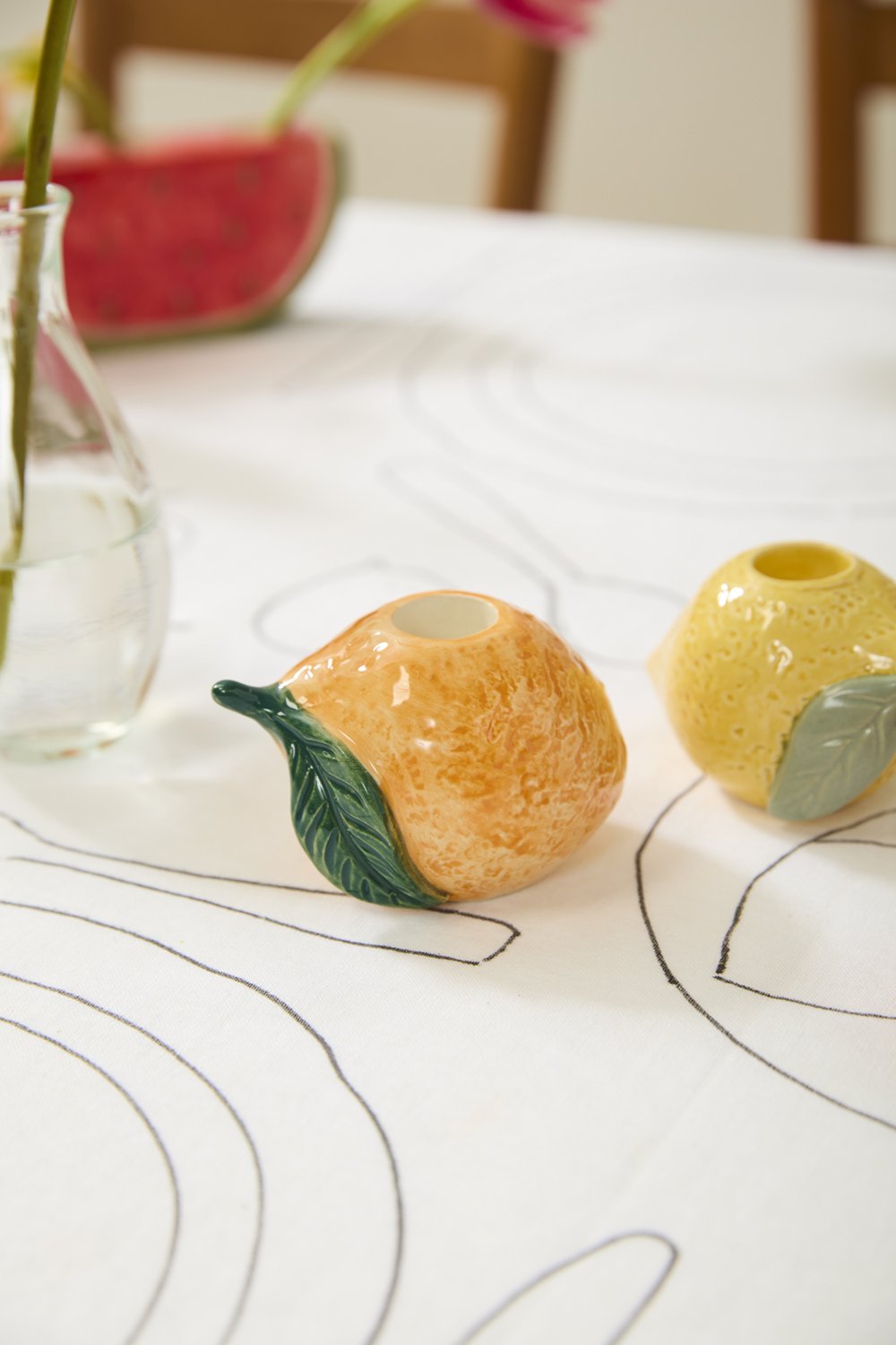 Orange candleholder on a white tablecloth beside a lemon candleholder and a glass bud vase