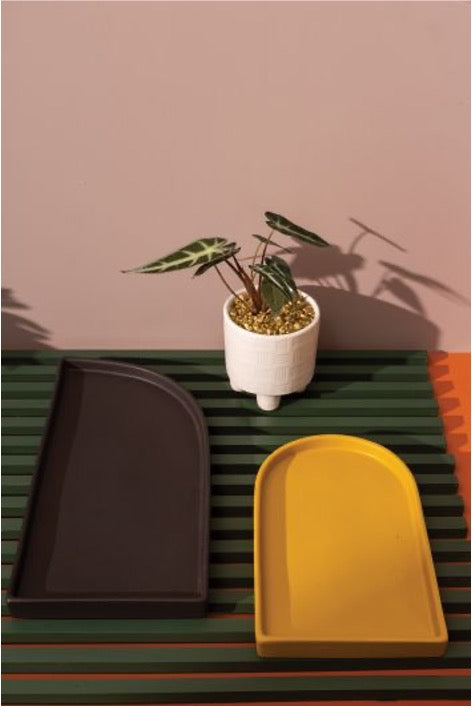 Set of ceramic trays. Arched tray in mustard and semi-arched tray in black on green slated table next to plant.