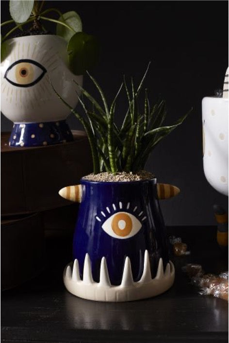 Monster teeth pot in blue with one eye in center, white teeth-like base with snake plant