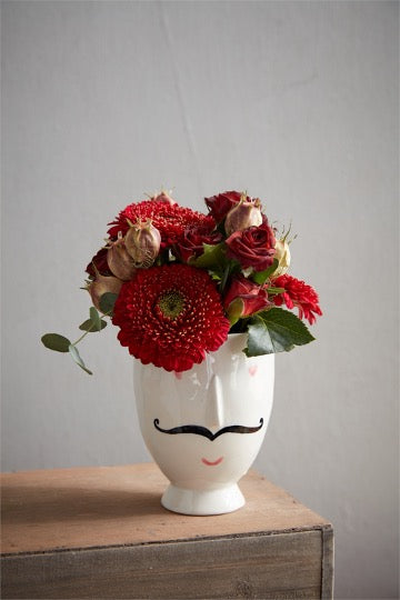 Footed white pot with pink heart eyes, black mustache, pink lips and raise nose. Red floral arrangement in pot.