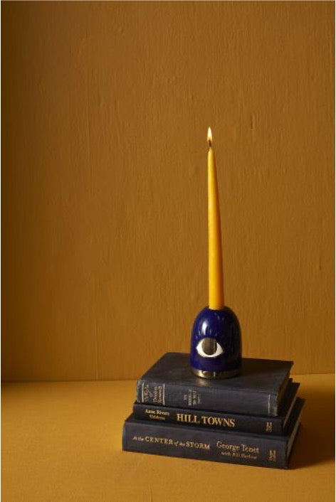 Candlestick holder in blue with eye in gold accents on stack of black book with lit orange taper candle.