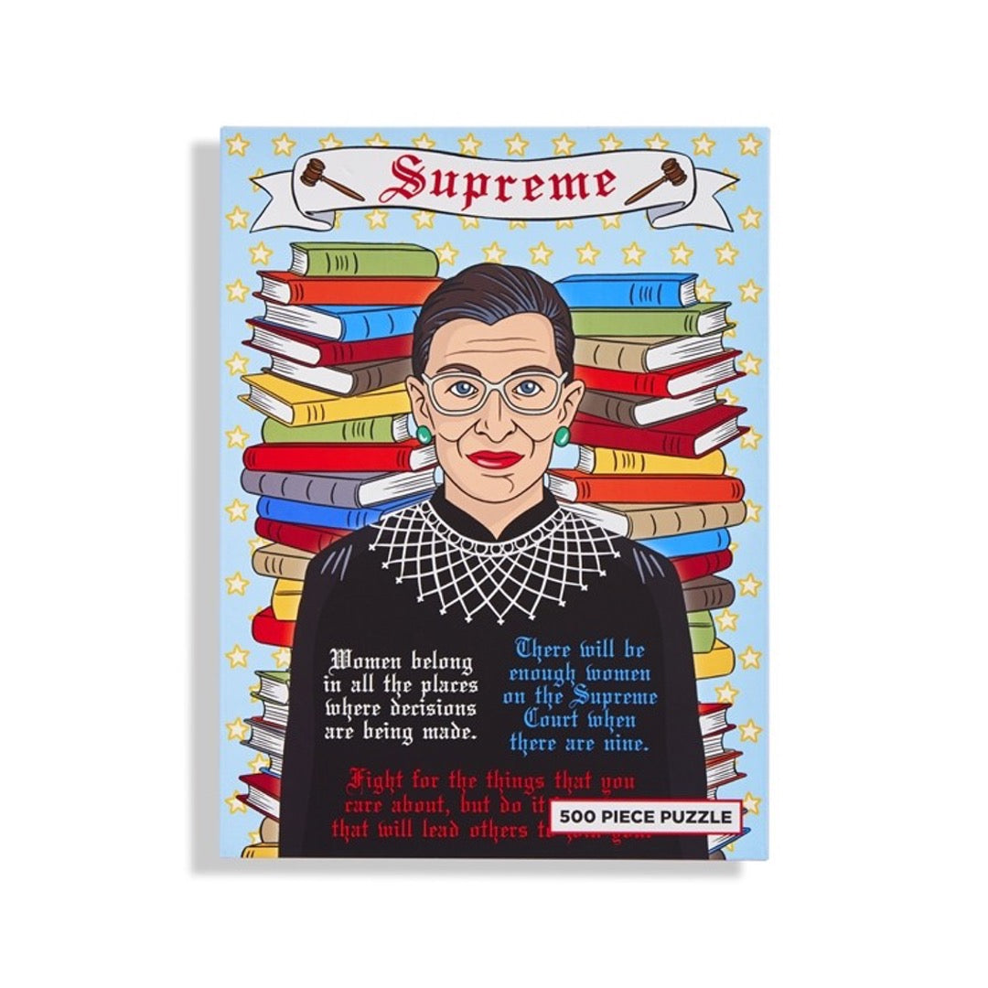 Front 500 piece puzzle box of Ruth Bader Ginsburg in front of stack of books with some of her famous quotes. Banner on top that says Supreme and 2 gavels on either side