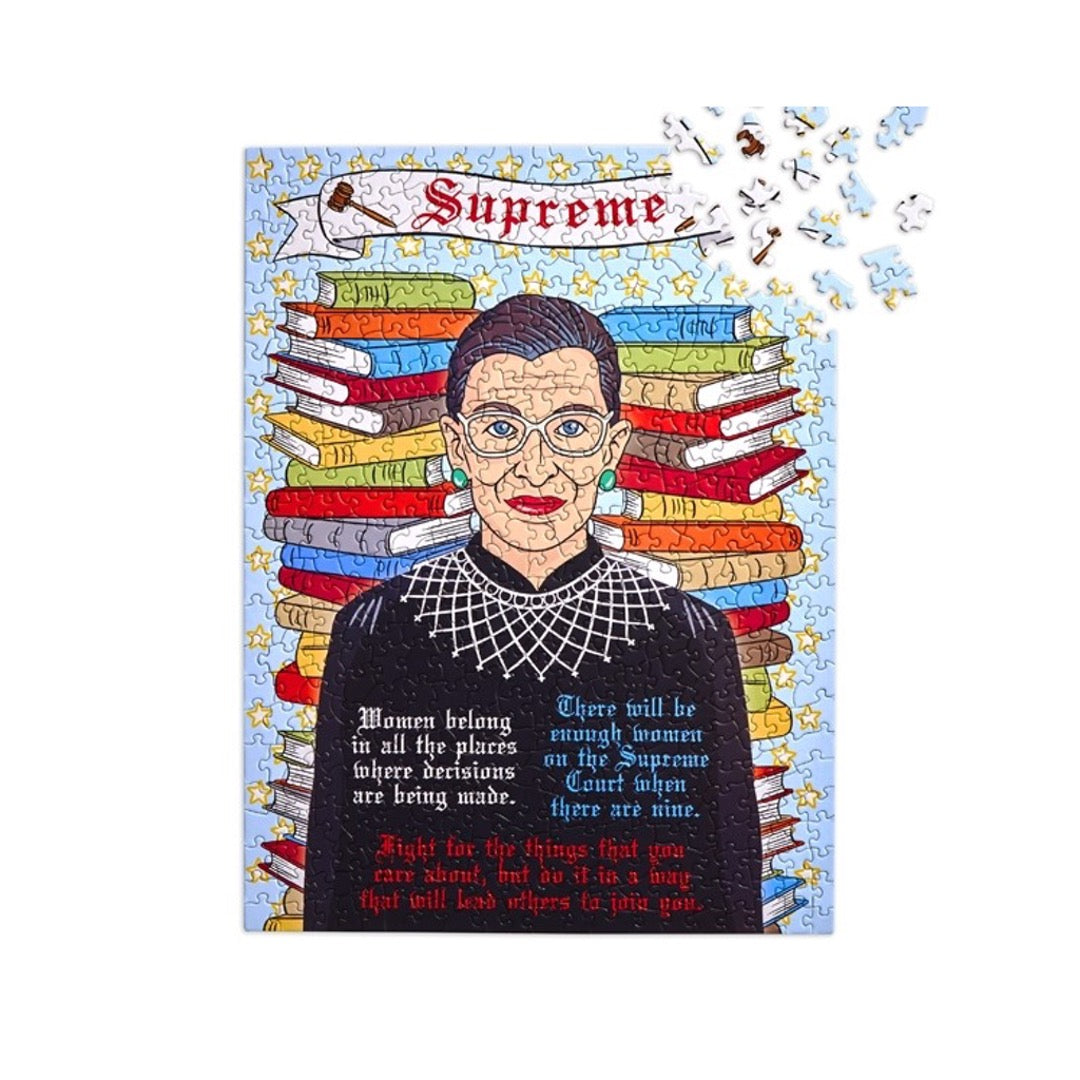 500 piece puzzle of Ruth Bader Ginsburg in front of stack of books with some of her famous quotes. Banner on top that says Supreme and 2 gavels on either side